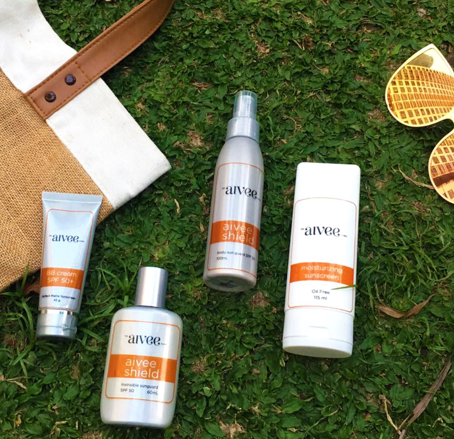 Aivee sunscreen products arranged on grass beside a straw bag and sunglasses